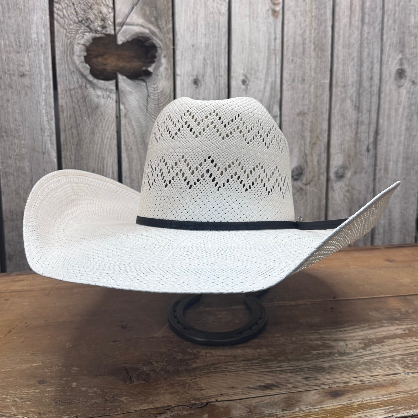 A white cowboy straw hat with a zig zag venting pattern across the crown, featuring a black leather sweatband and a black cord ribbon band, displayed against a wooden background.