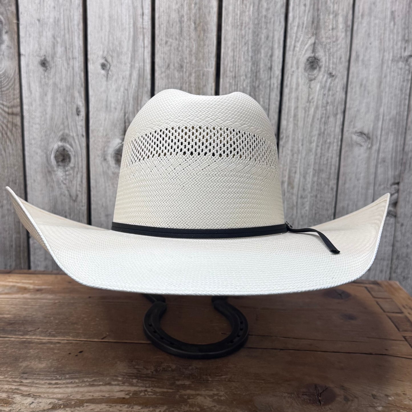A white straw cowboy hat with a 4 1/2 inch brim, featuring a black leather sweatband and a black cord ribbon band, displayed against a wooden background.