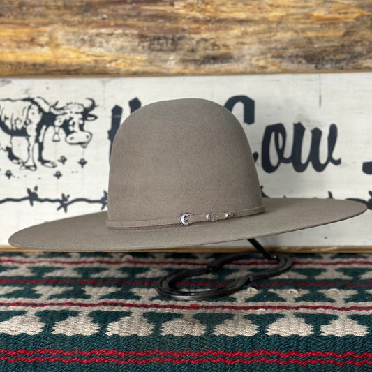 A beige-colored, 7X premium pecan-colored felt hat with a 5-inch brim, featuring a sweatband and a buckle on the band, displayed on a surface with a patterned rug and a sign in the background.