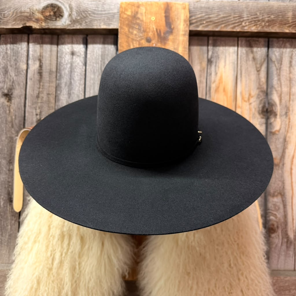 A black wide-brimmed felt hat displayed on a cream-colored faux fur stand against a rustic wooden plank background.