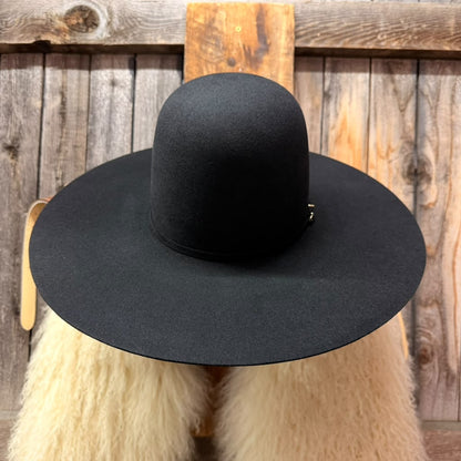 A black wide-brimmed felt hat displayed on a cream-colored faux fur stand against a rustic wooden plank background.