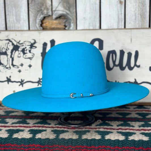 A turquoise blue Rodeo King cowboy hat with an open crown, hand-shaped brim, and a metal buckle on the band, displayed against a background with a cow print.