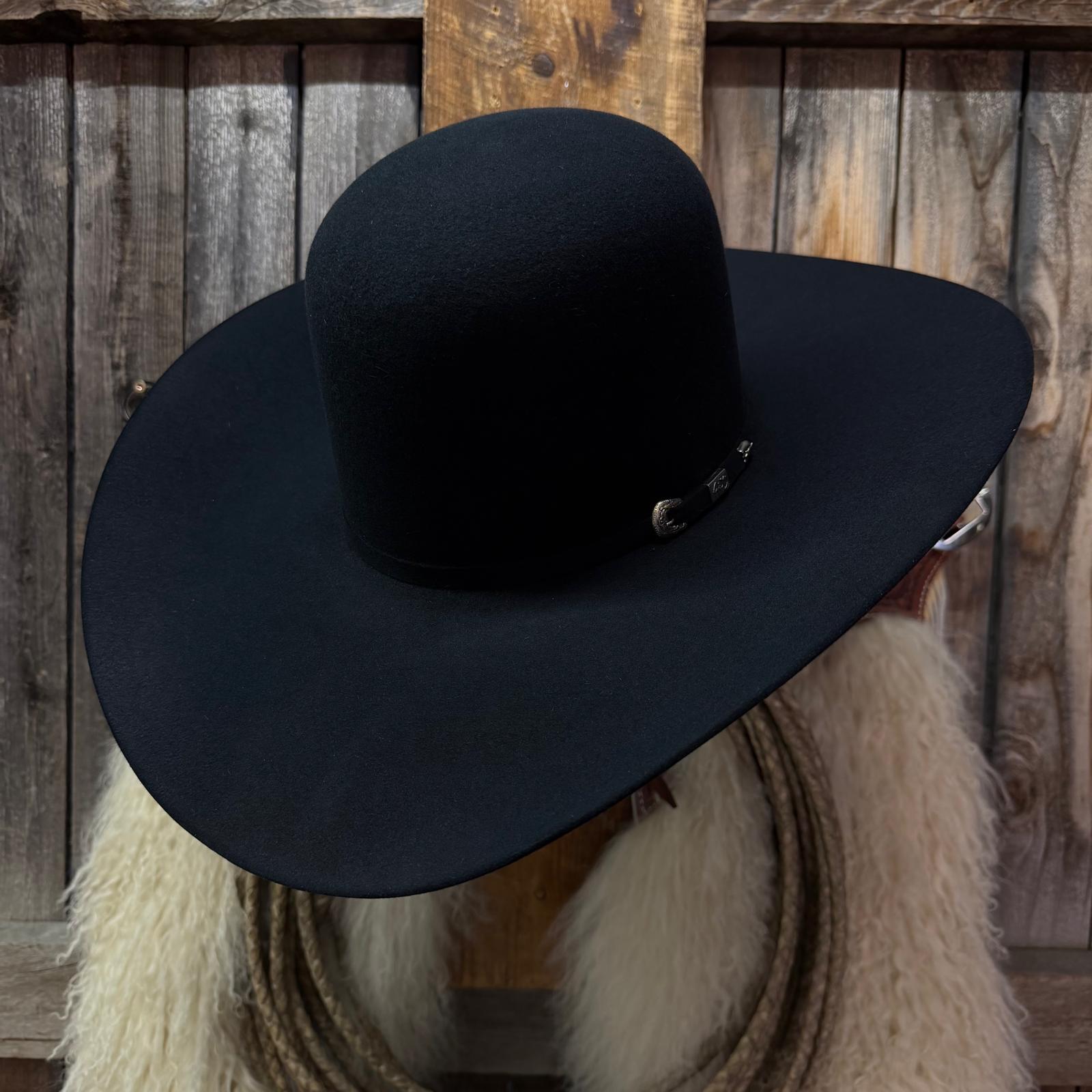 A black felt cowboy hat with a small silver buckle detail, displayed on a mannequin head with a white faux fur collar in front of a rustic wooden plank background.