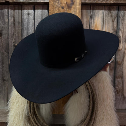 A black felt cowboy hat with a small silver buckle detail, displayed on a mannequin head with a white faux fur collar in front of a rustic wooden plank background.
