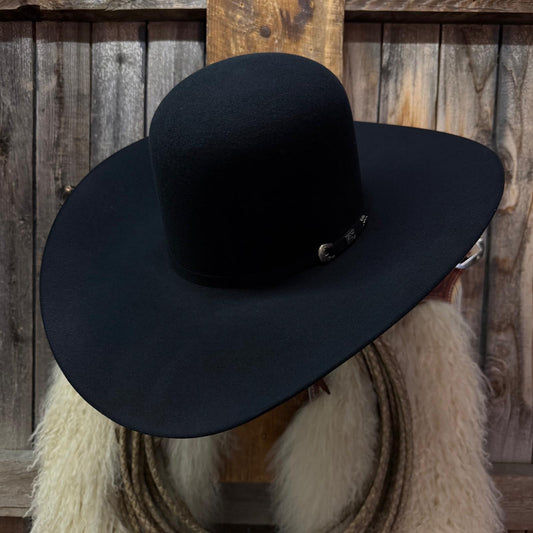 A black felt cowboy hat with a small silver buckle detail, displayed on a mannequin head with a white faux fur collar in front of a rustic wooden plank background.