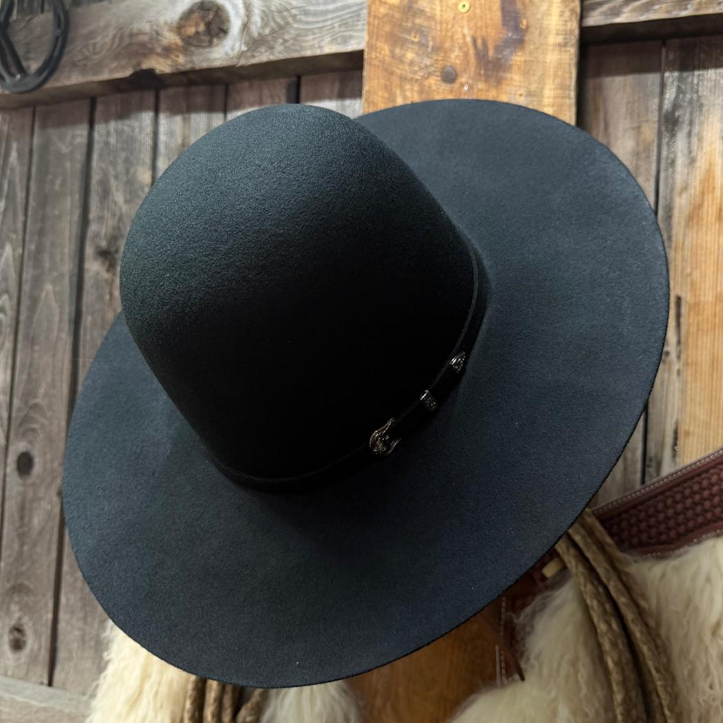 A black wide-brim fedora hat displayed on a wooden stand in front of a rustic wood plank wall, with a white fur trim and rope visible below.