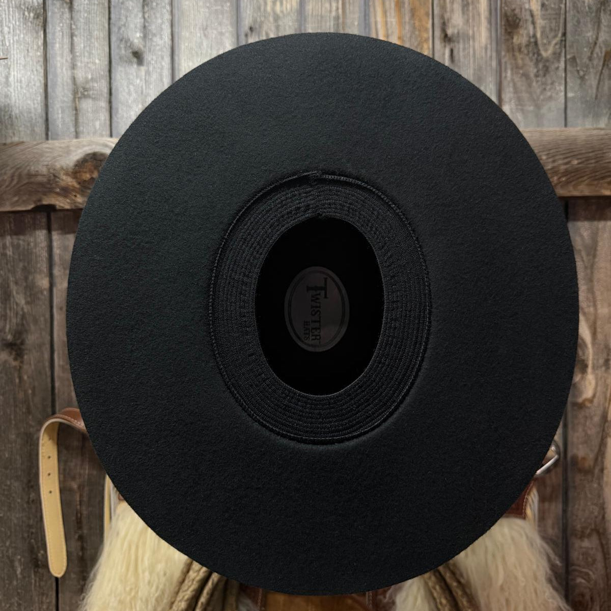 A black wide-brim felt cowboy hat shown from the back, featuring a circular center opening with a visible 'Twister' brand label, set against a rustic wooden plank background.