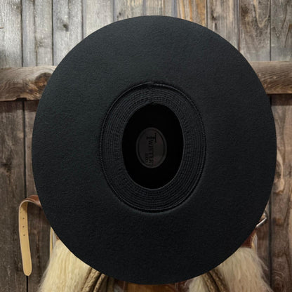 A black wide-brim felt cowboy hat shown from the back, featuring a circular center opening with a visible 'Twister' brand label, set against a rustic wooden plank background.