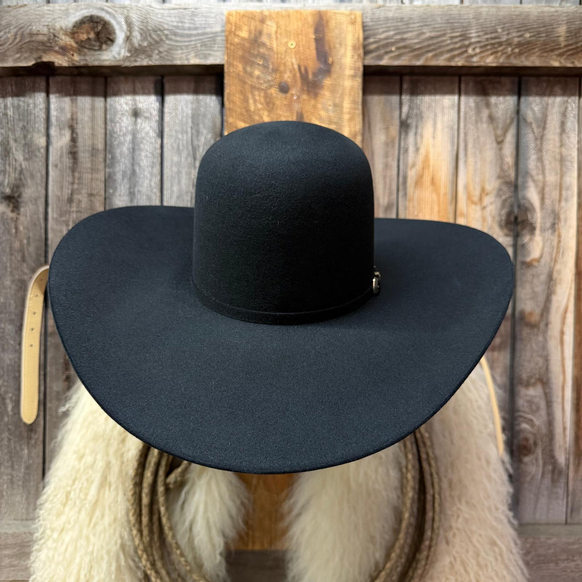 A black wide-brim cowboy hat displayed on a rustic wooden stand with a white fur collar and rope in the background.