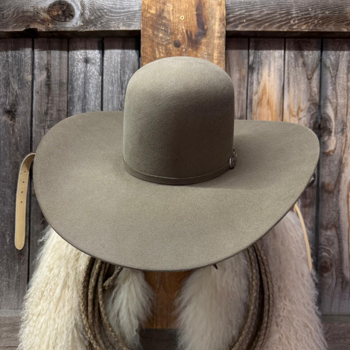 A wide-brimmed felt cowboy hat in a muted taupe-brown color, displayed on a rustic wooden stand with a white fur collar and rope against a weathered wood plank background.
